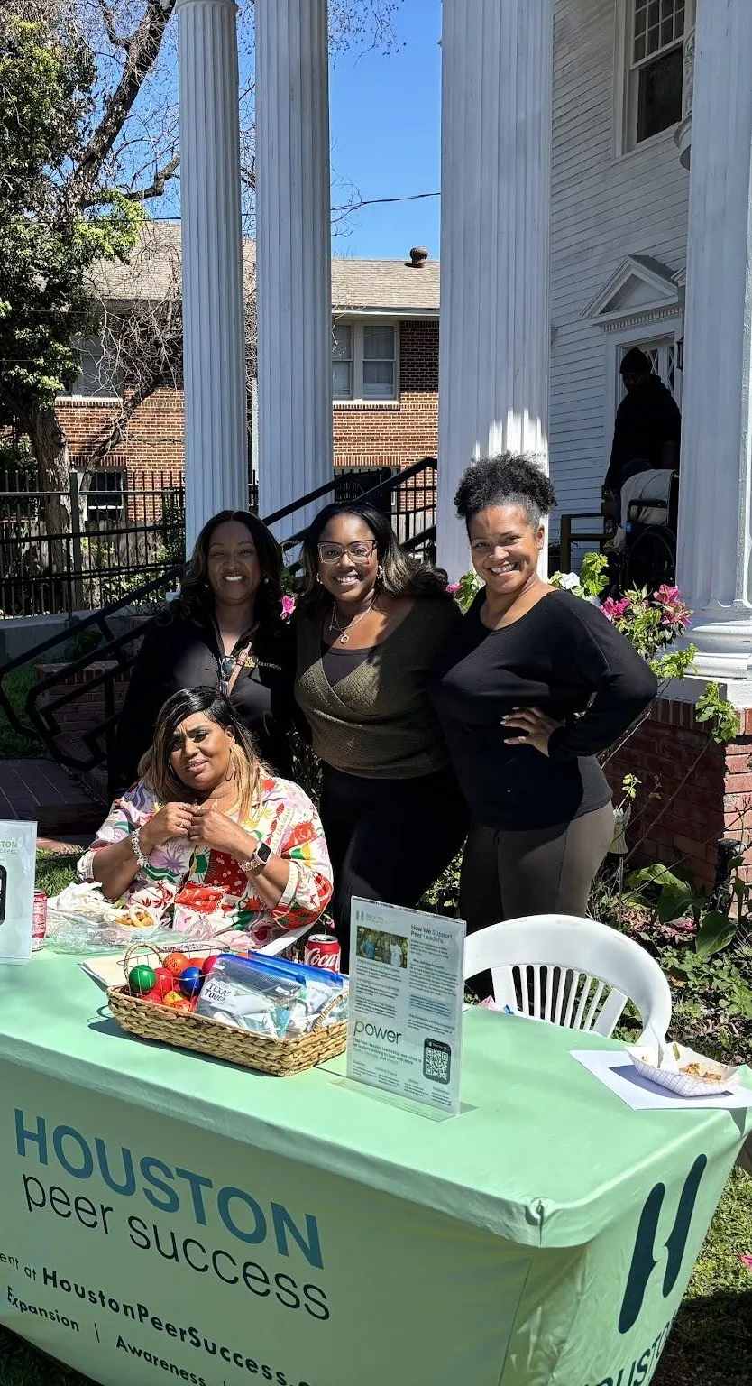 Cuatro mujeres de Houston Peer Success sonriendo detrás de su mesa informativa verde menta, afuera de una casa blanca con columnas