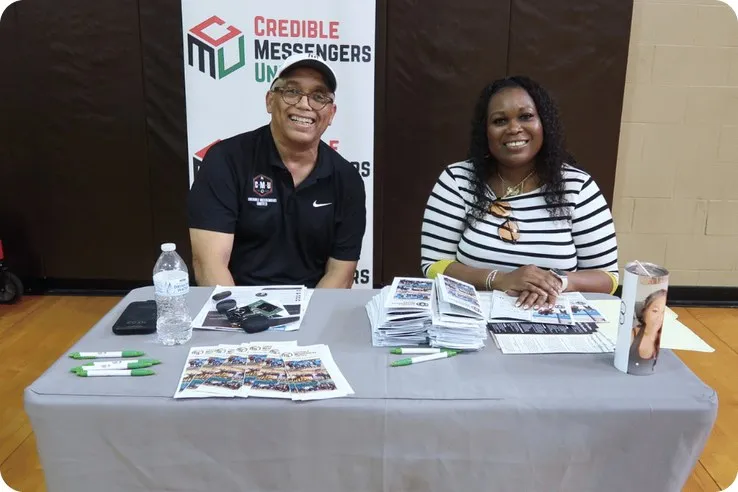 Dos representantes de Credible Messengers United sonriendo en su mesa durante una feria comunitaria de recursos