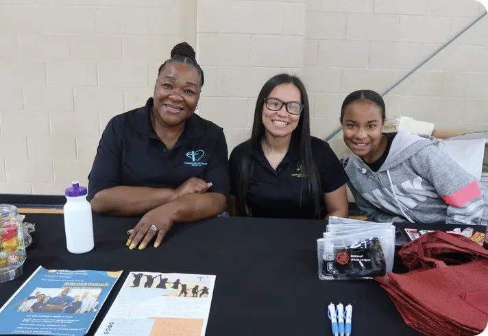 Tres mujeres sonrientes detrás de una mesa de información en un evento comunitario de Houston Peer Success