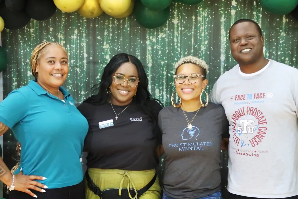 Cuatro personas sonrientes, abrazadas frente a un arco de globos verdes y dorados en un evento comunitario